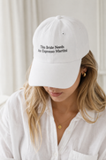 A woman wearing a white baseball cap with black embroidered text that reads 'This Bride Needs An Espresso Martini'. The photo is taken in a bright, soft-focus indoor setting.