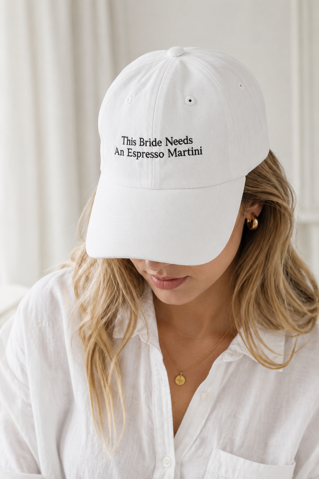 A woman wearing a white baseball cap with black embroidered text that reads 'This Bride Needs An Espresso Martini'. The photo is taken in a bright, soft-focus indoor setting.