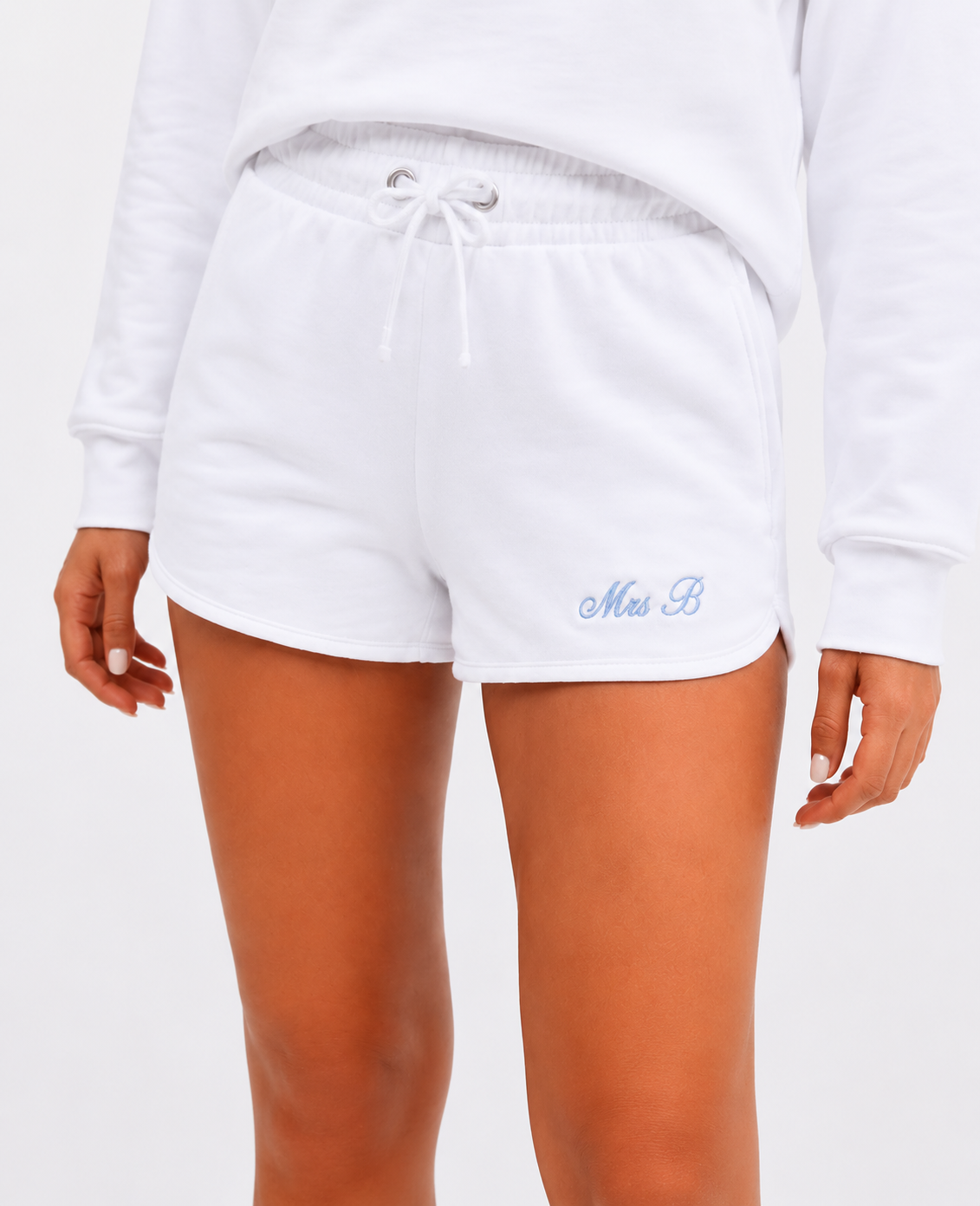 White sweat shorts with a drawstring waist and a light blue embroidered 'Miss B' logo on the left leg, shown on a model wearing a white sweatshirt.