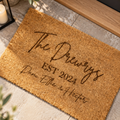 A personalized coir doormat with the text 'The Drewrys EST 2024 Dane, Ellie & Hector' printed in dark brown script, shown on a stone tile floor with a candle and wooden trim nearby.