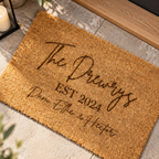 A personalized coir doormat with the text 'The Drewrys EST 2024 Dane, Ellie & Hector' printed in dark brown script, shown on a stone tile floor with a candle and wooden trim nearby.