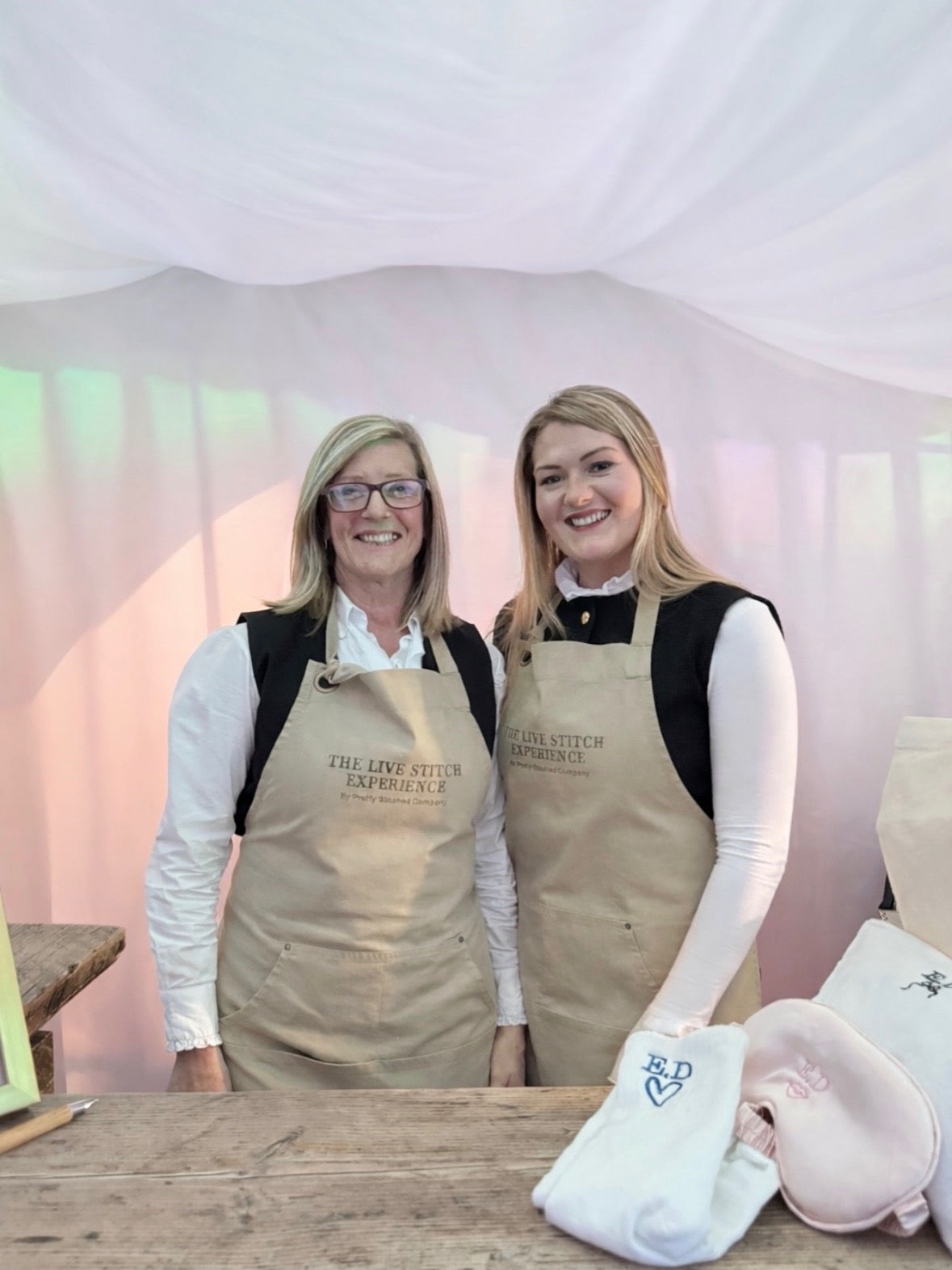 Two women wearing beige aprons with 'The Live Stitch Creamery' logo stand behind a wooden table under a white canopy tent. A folded white towel with a blue embroidered heart and a pink hat are on the table in the foreground.